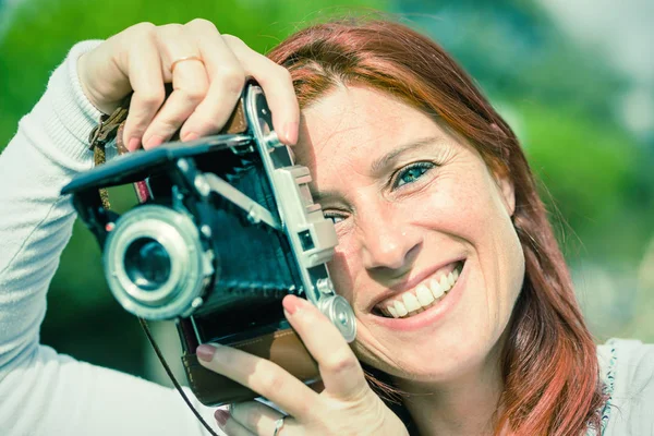Retrato de una mujer pelirroja muy sonriente tomando fotos con una cámara vieja . — Foto de Stock