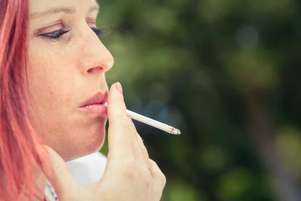 Portrait rapproché d'une jolie femme rousse crachée de fumée de cigarette dans un parc avec de la verdure en arrière-plan — Photo