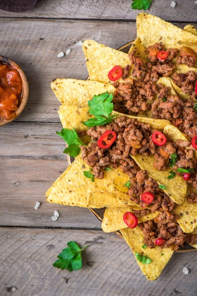 Mexican nacho corn tortilla chips with cheese, meat, guacamole and red hot spicy salsa. Nachos with ground beef on wooden background, copy space.