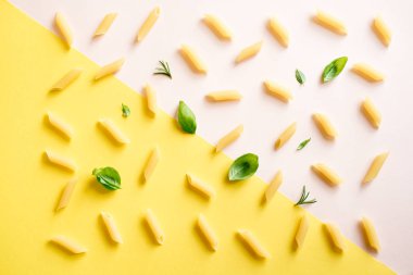 Penne makarna pembe pastel ve sarı arka plan, üstten görünüm, düz yatıyordu. Pişmemiş penne makarna otlar fesleğen ve biberiye ile desen.