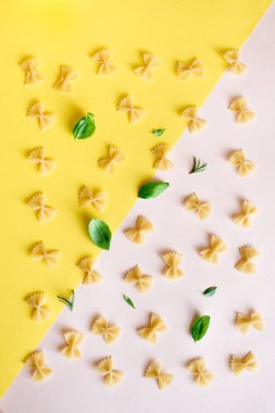 Farfalle makarna üzerine pembe pastel ve sarı arka plan, üstten görünüm, düz yatıyordu. Pişmemiş farfalle makarna otlar fesleğen ve biberiye ile desen.