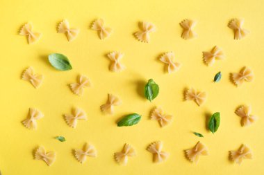 Farfalle makarna sarı arka plan üzerine üst görünüm, düz yatıyordu. Pişmemiş farfalle makarna fesleğen yaprakları ile desen.
