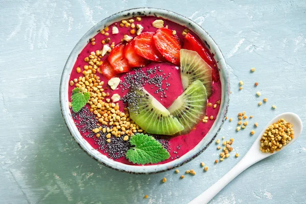 Acai smoothie bowl with chia seeds, fruits, berries (strawberries) and bee pollen for healthy vegan vegetarian diet raw breakfast. Breakfast smoothie bowl on blue background.