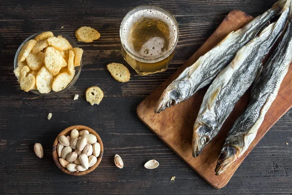 Assorted snacks for beer: sun dried fish, nuts, salted croutons or crackers over wooden background, top view, copy space. Lager beer and snacks with dried smelts.