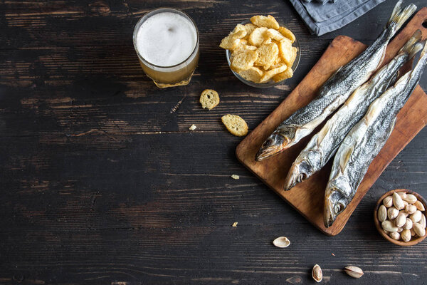 Assorted snacks for beer: sun dried fish, nuts, salted croutons or crackers over wooden background, top view, copy space. Lager beer and snacks, dried smelts.