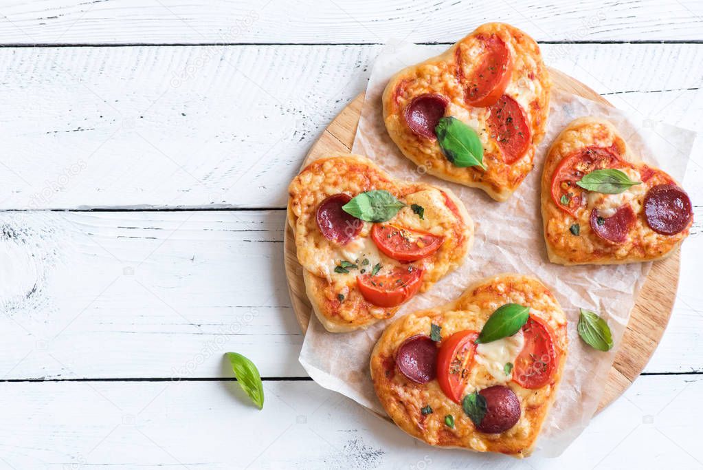 Heart shaped mini pizzas over wooden background with copy space. Mini pizzas with tomatoes, pepperoni, mozzarella cheese and basil for Valentine's day, love concept.