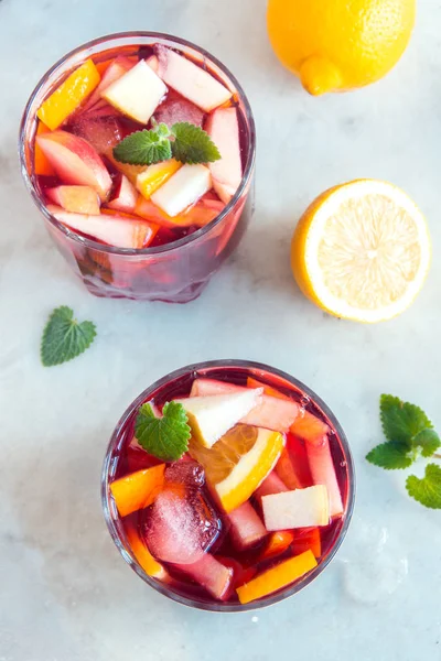 Red wine sangria or punch with fruits, mint and ice in glasses. Homemade refreshing fruit sangria over rustic white table, copy space