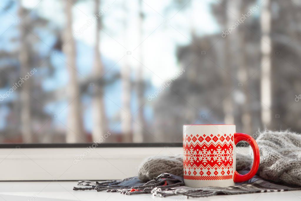 Red cup of hot drink on the window sill, with winter landscape in snow. Cozy home, winter weekend and holidays, Christmas morning concept.