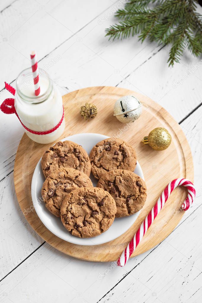 Chocolate chip cookies and Milk for Santa. Traditional Christmas homemade cookies, candy cane and bottle of milk with Christmas decor on white wooden table, top view.