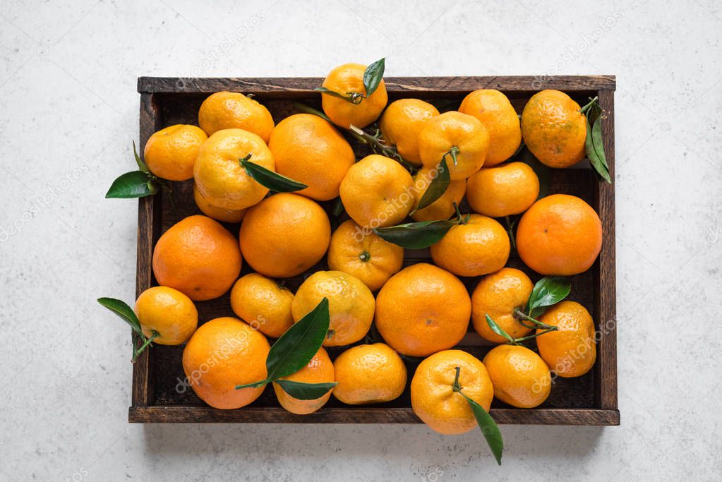 Tangerines (oranges, clementines, citrus fruits) with green leaves in box on white stone background, top view.