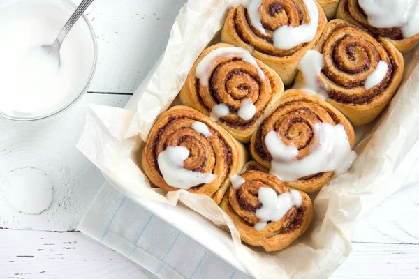 Cinnamon rolls or cinnabon, homemade recipe of sweet traditional dessert buns with white cream sauce on white wooden background.