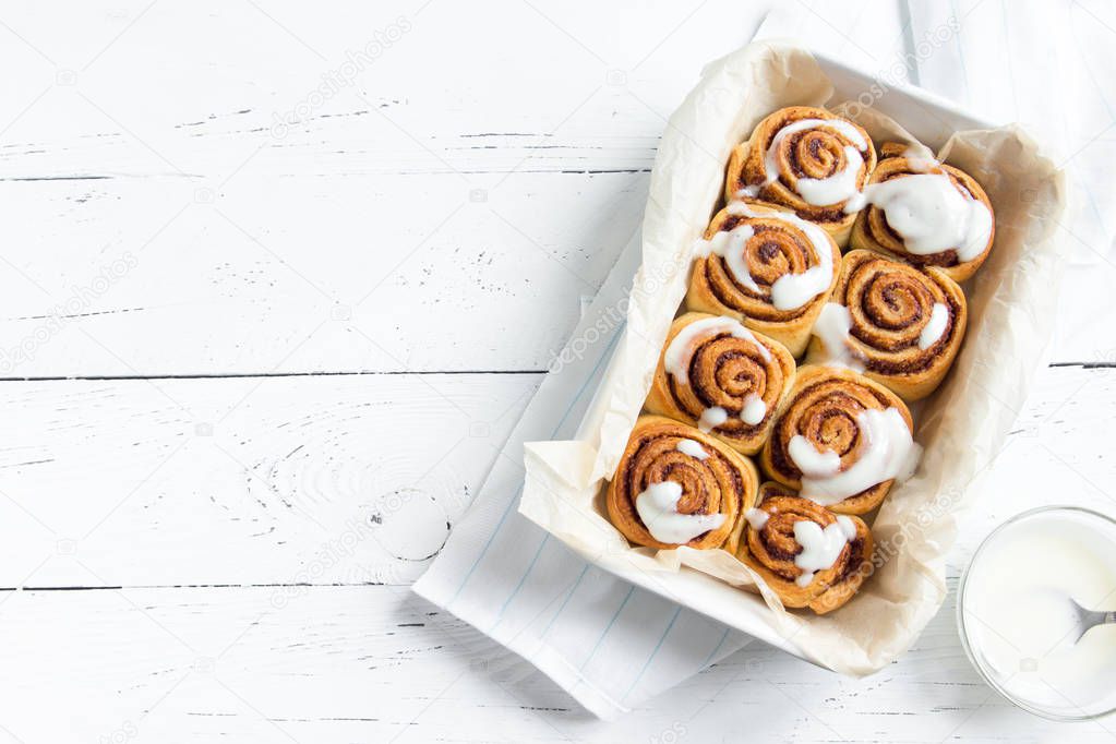 Cinnamon rolls or cinnabon, homemade sweet traditional dessert buns with white cream sauce on white wooden background, copy space.