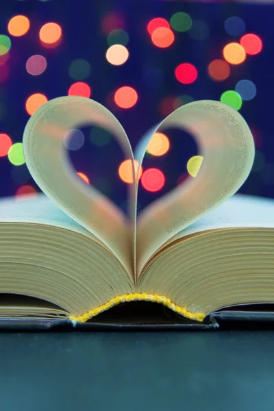 Open book with heart shape from paper pages on table in soft night light holiday style close up with bokeh blur background . for christmas and valentine\'s day. Copy space, front view.