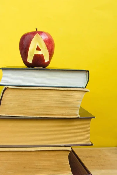 Back to school and knowledge concept. Stack of books and fresh red apple with mark A on wooden background and trendy yellow wall. Front view