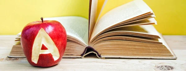 Back to school and knowledge concept. Stack of books and fresh apple with mark A on wooden background and trendy yellow wall. Banner