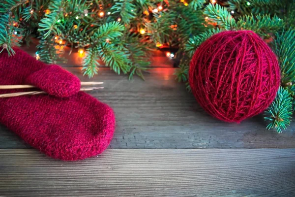 Red wool mitten with yarn ball and wooden knitting needles near green christmas fur tree and bright colorful lights on rustic background. Toned. Christmas card, postcard concept. Front view