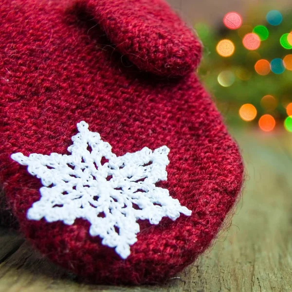 Red wool mitten with white cotton crocheted snowflake on the rustic wooden background. Red, blue, yellow and green spots of christmas lights. on fur tree. Card, postcard concept. Front view.Bokeh.
