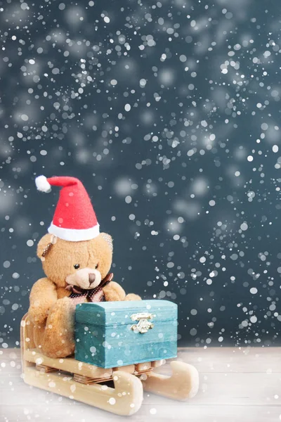 Decoration Santa's helper bear in a red hat rides a sleigh with a chest of gifts in the snowfall at Christmas. Front view, close up. Snowflake. Holiday greeting card concept. Copy space