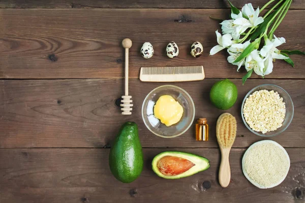 Ingredients for vitamin avocado skin care facial mask green lime, bottle cosmetic oil,honey,dipper, oat-flakes, hair brush on brown wooden background. Alstroemeria flower. Top view close up copy space