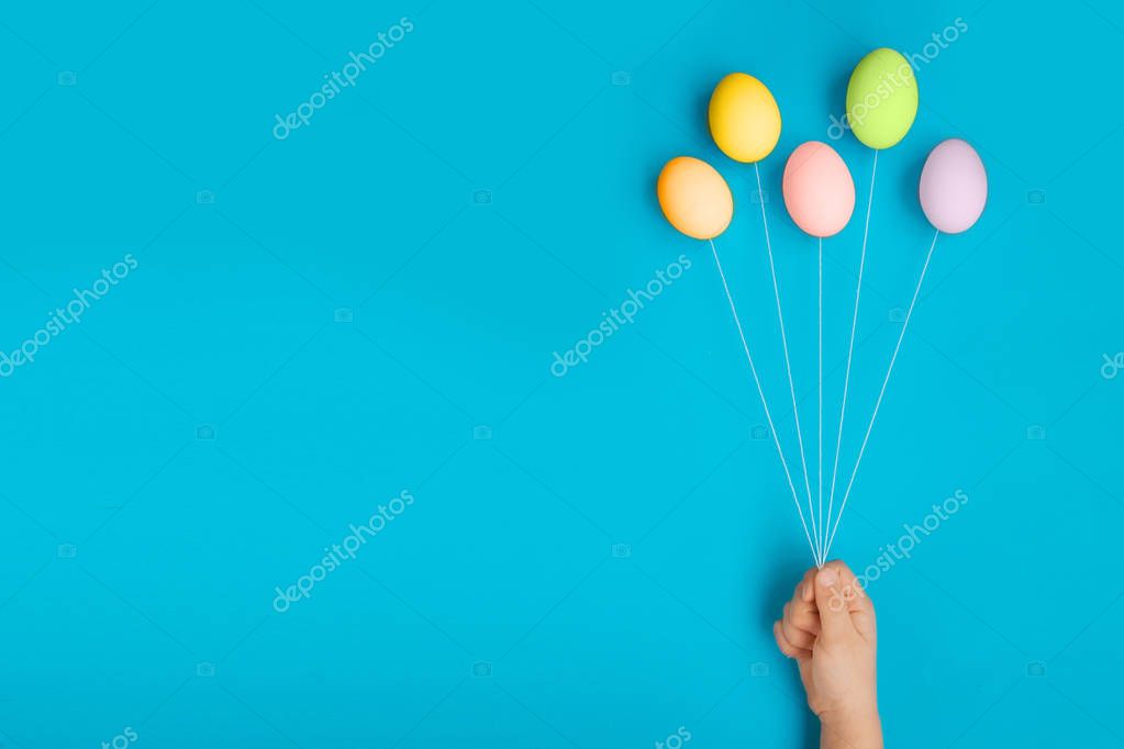 Beautiful creative decoration background idea with child's hand holding ropes flying balloons painted colorful eggs on bright blue paper. Vibrant spring card concept. Close up.Front view. Copy space.