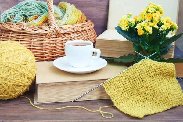 Knitting of yellow wool with needles, white espresso coffee cup saucer, basket, old books, Kalanchoe on wooden rustic background. Front view