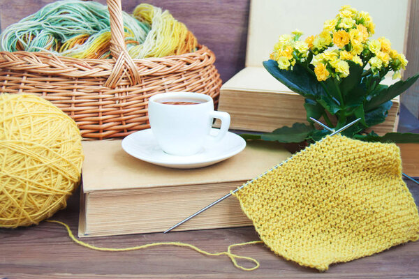 Knitting of yellow wool with needles, white espresso coffee cup saucer, basket, old books, Kalanchoe on wooden rustic background. Front view