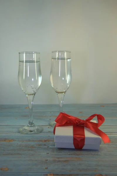 Two glasses with champagne and gift box with satin ribbon on a wooden table. Close-up.