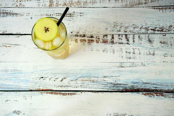 A tall glass of apple juice with ice stands on a wooden table next to green apples. Flat lay.