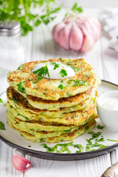 Zucchini pancakes with parsley and sour cream, summer food, delicious snack. High stack in a plate on white wood