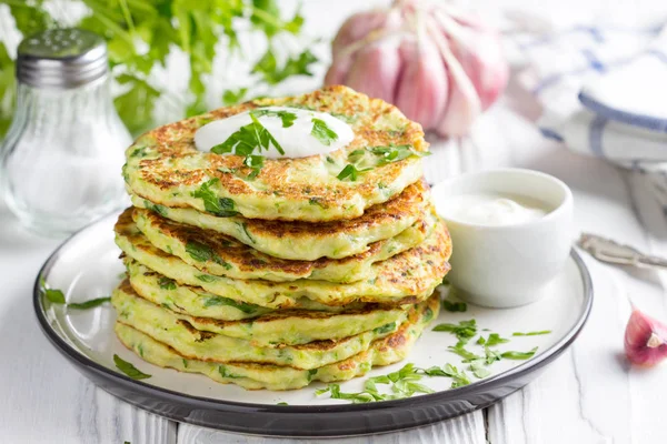 Zucchini pancakes with parsley and sour cream, summer food, delicious snack. High stack in a plate on white wood