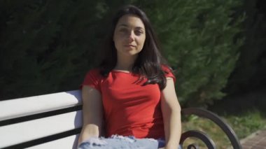 Kamerada poz veren güzel kız. Yeşil gözlü, kırmızı tişörtlü, bankta oturan genç ve güzel bir kız.