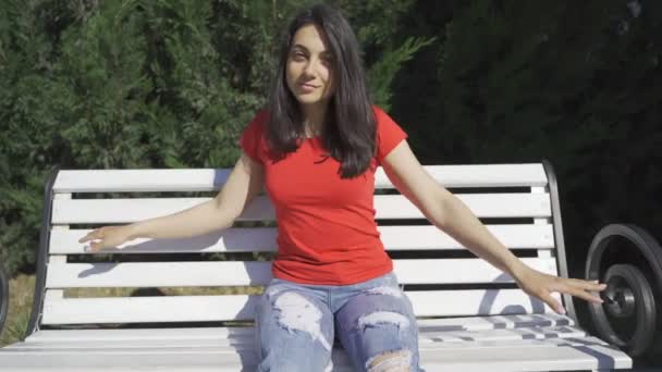 예쁜 여자가 카메라에 찍혔네. 붉은 티셔츠를 입고 벤치에 앉아 있는 녹색 눈의 아름다운 소녀 — 비디오