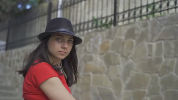 Pretty girl posing on camera. Young beautiful girl with green eyes in a red T-shirt and black hat posing on camera in the summer — Stock Video
