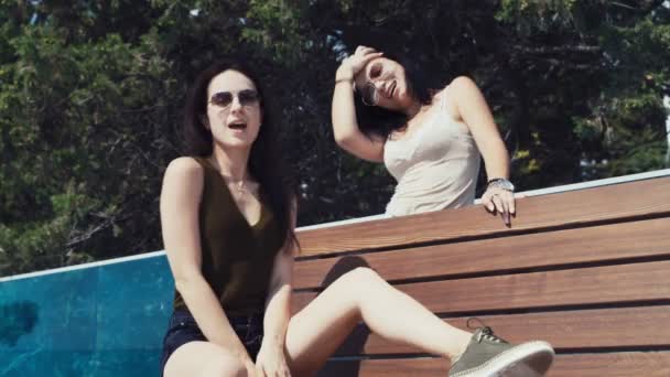 Két gyönyörű barna lány napszemüvegben táncol állva és ülve egy padon tenger — Stock videók