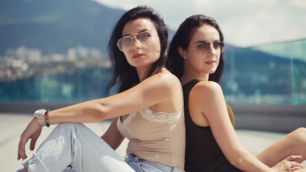Δύο Όμορφα Καστανά Κορίτσια Γυαλιά Ηλίου Κάθονται Πλάτη Πλάτη Χαμογελώντας — Αρχείο Βίντεο