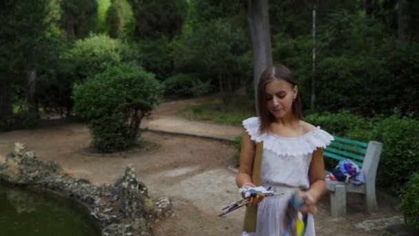 Menina artista prepara paleta de papel e pincéis de pintura para pintura de paisagem no parque com uma pequena lagoa 4k — Vídeo de Stock