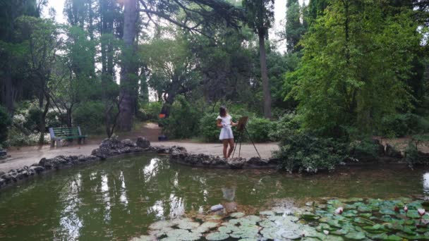 Chica artista en el parque pinta un paisaje con pinturas al óleo un pequeño estanque viejo 4k — Vídeos de Stock