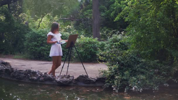 Chica artista en el parque pinta un paisaje con pinturas al óleo un pequeño estanque viejo 4k — Vídeo de stock