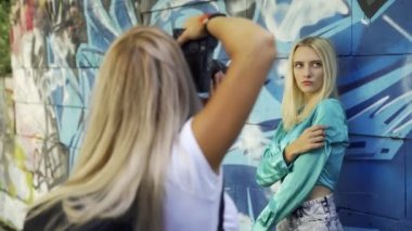 Sarışın kız şehirde yaz aylarında graphitti ile duvarda bir kız fotoğrafçı için poz veriyor. Güzel genç kız modeli bir kız fotoğrafçı için poz. Grafiti duvarı. 4k