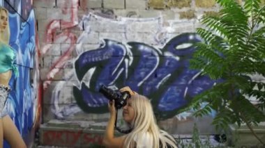 Sarışın kız şehirde yaz aylarında graphitti ile duvarda bir kız fotoğrafçı için poz veriyor. Güzel genç kız modeli bir kız fotoğrafçı için poz. Grafiti duvarı. 4k