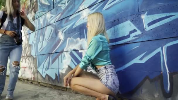 ブロンドの女の子は 市内の夏にグラフィッティと壁に女の子の写真家のためにポーズ 美少女モデルが女の子写真家のポーズ 落書きの壁4K — ストック動画
