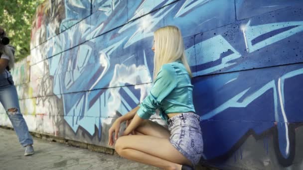 ブロンドの女の子は 市内の夏にグラフィッティと壁に女の子の写真家のためにポーズ 美少女モデルが女の子写真家のポーズ 落書きの壁4K — ストック動画