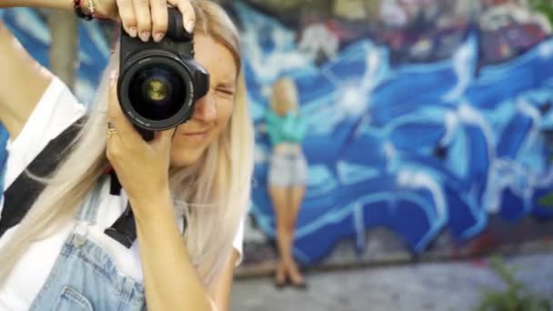 在夏天 金发女孩在墙边为一位女摄影师摆姿势 美丽的年轻女孩模型冒充一个女孩摄影师 涂鸦墙 — 图库视频影像