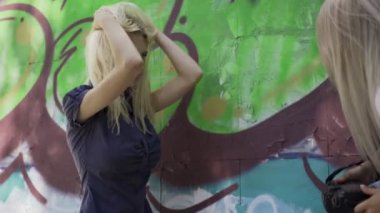 Sarışın kız şehirde yaz aylarında graphitti ile duvarda bir kız fotoğrafçı için poz veriyor. Güzel genç kız modeli bir kız fotoğrafçı için poz. Grafiti duvarı. 4k