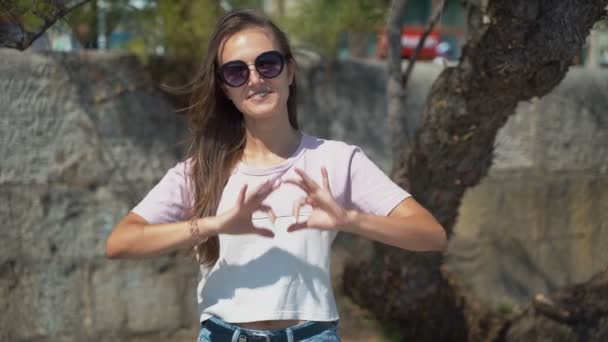 Hermosa chica muestra un corazón con las manos. Joven morenita hermosa en gafas de sol con sus manos muestra un corazón. — Vídeos de Stock