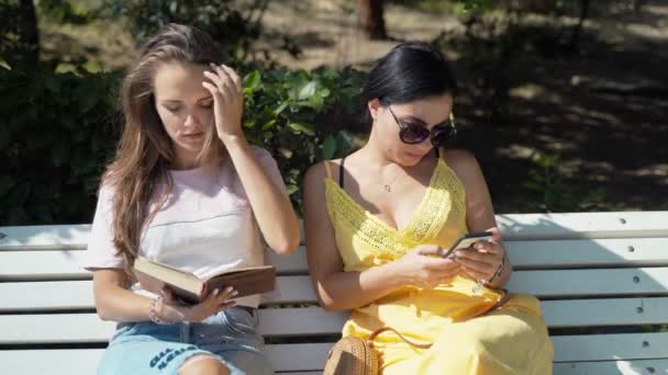 Dos chicas jóvenes miran un teléfono móvil y luego comienzan a leer un libro . — Vídeos de Stock