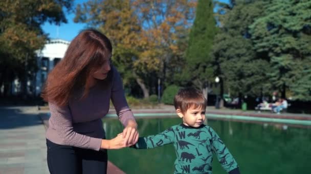 Μια νεαρή μητέρα με ένα γιο δύο ετών περπατά στην πόλη κοντά στη λίμνη. Ηλιόλουστη ζεστή μέρα το φθινόπωρο. Ένα μικρό αγόρι περπατά με τη μητέρα του στην πόλη, χαμογελάει και χαίρεται. — Αρχείο Βίντεο