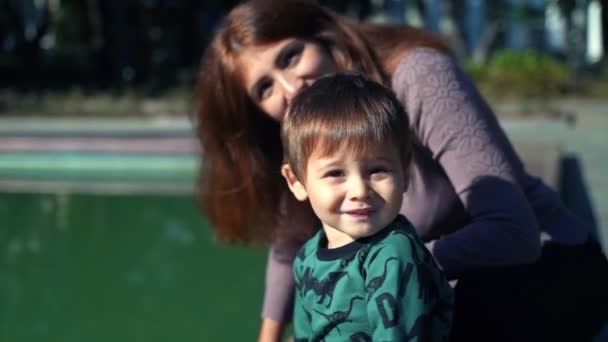 Μια νεαρή μητέρα με ένα γιο δύο ετών περπατά στην πόλη κοντά στη λίμνη. Ηλιόλουστη ζεστή μέρα το φθινόπωρο. Ένα μικρό αγόρι περπατά με τη μητέρα του στην πόλη, χαμογελάει και χαίρεται. — Αρχείο Βίντεο