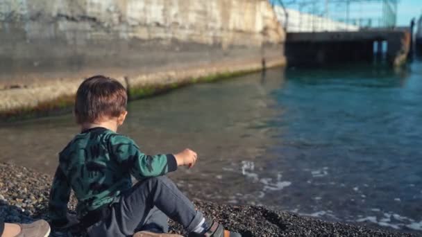 Eine junge Mutter und ihr Sohn gehen in der Stadt am Meer spazieren und werfen Steine ins Wasser. Ein kleiner Junge geht mit seiner Mutter ans Meer und wirft an einem sonnigen Tag Steine ins Wasser. — Stockvideo