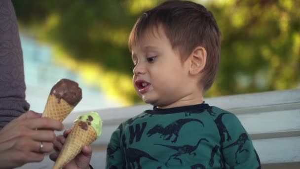 息子と一緒にアイスクリームを食べるお母さん。ママと子供子供はアイスクリームを食べる。ベンチにいる母親と小さな男の子はアイスクリームを食べていますが、子供は母親とアイスクリームを交換したいと思っています — ストック動画